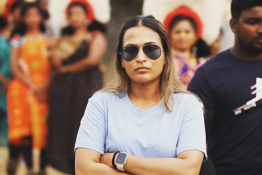 Aishwarya Rajinikanth on a film set, wearing sunglasses with her arms folded, crew and background artistes behind her