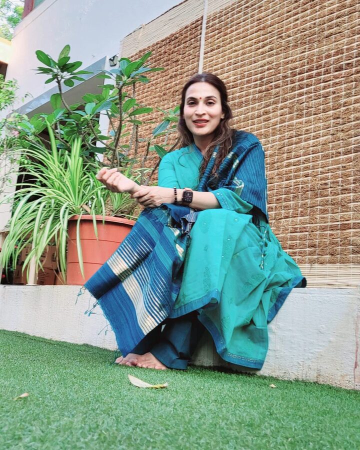 Aishwarya Rajinikanth seated outdoors in a teal handloom saree against a brick wall